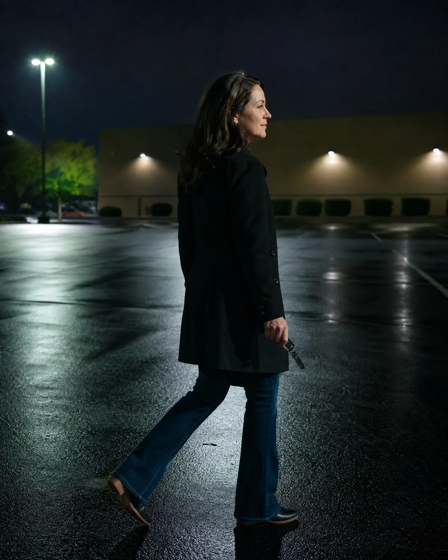 A woman in a long dark coat and jeans walks across a wet, empty parking lot at night, holding keys, with a low lit building and a tall light pole behind her.